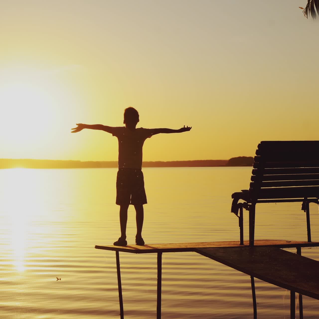 Boy doing yoga exercises by the river at sunset. Silhouette of a boy on an old bridge by the river. Beautiful summer sunset