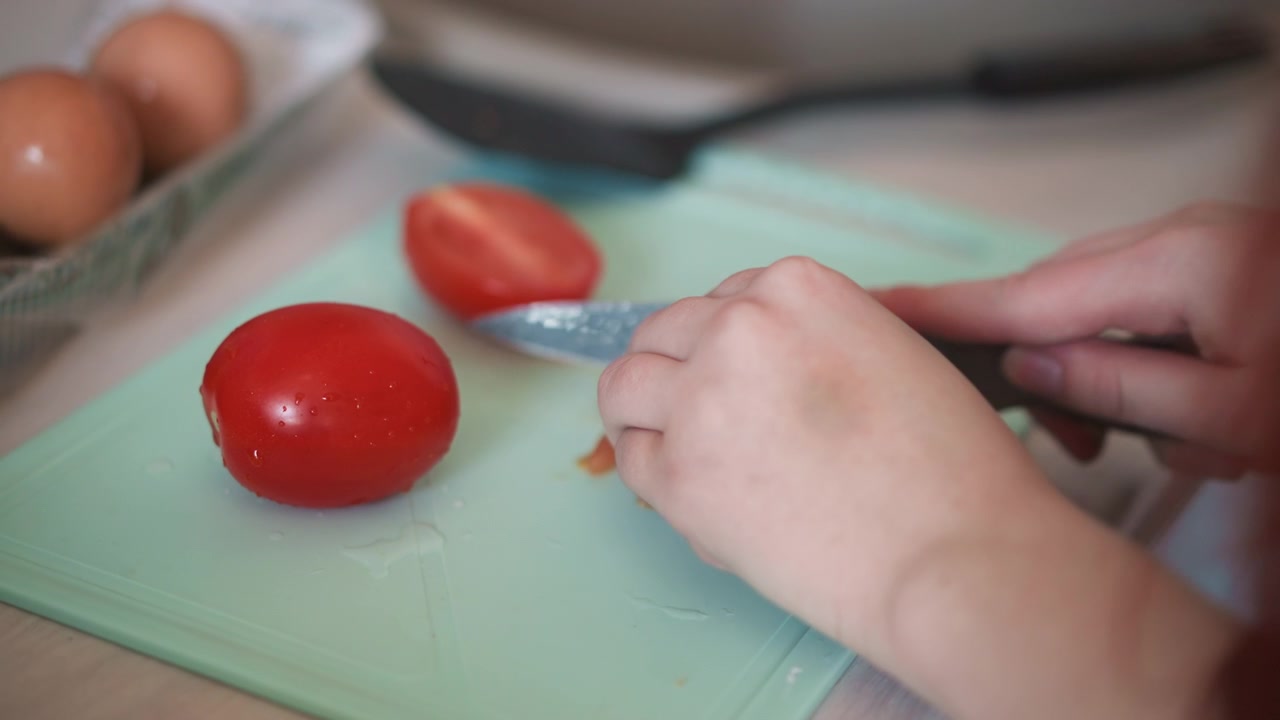 Primo piano di mani che affettano un pomodoro maturo su un tagliere con uova e pane nelle vicinanze, luce soffusa del mattino, preparazione calma e concentrata per la colazione, cuoco principiante che pratica le abilità con il coltello, polpa e succo rosso vibrante.