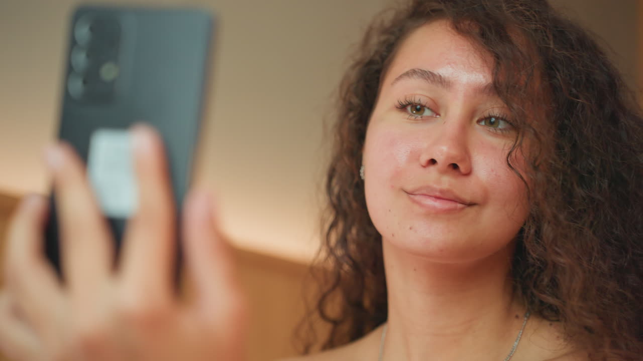 Donna multietnica in camera d'albergo che si applica il mascara mentre controlla il telefono, con una luce calda dal comodino. Espressioni animate, atmosfera di viaggio informale, energia per la creazione di contenuti e una fiducia rilassata.
