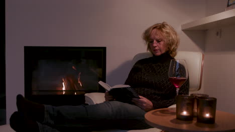 woman sitting by the fireplace reading a book and drinking a glass of wine