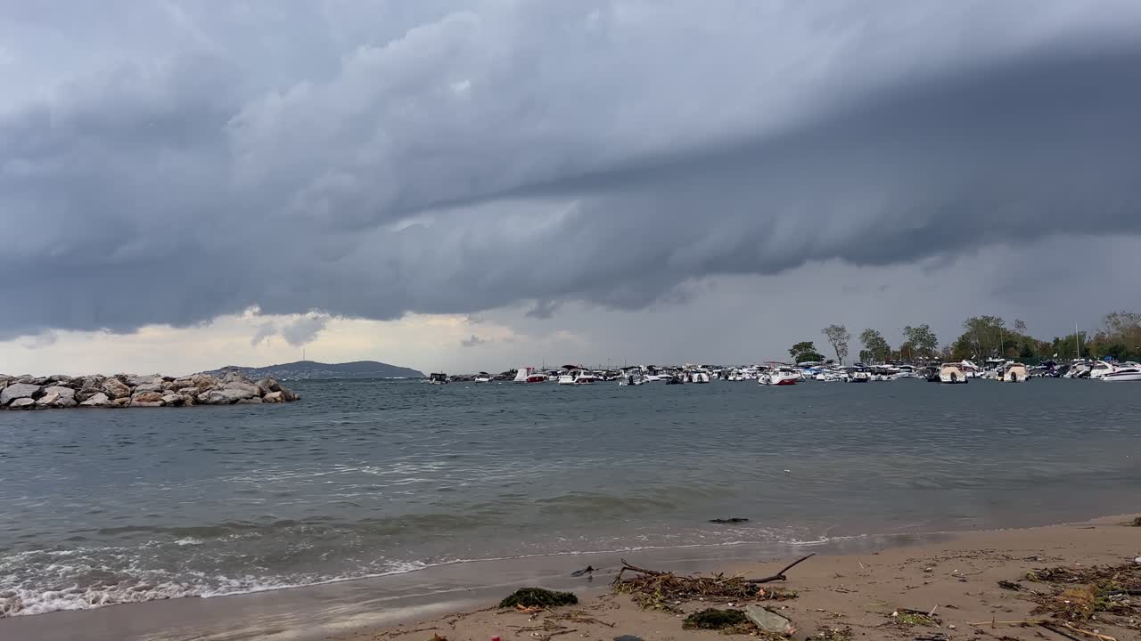 Dramatic dark storm clouds gather over a rough sea. Waves crash on the shore near a stone breakwater and a marina with boats on a gloomy autumn day