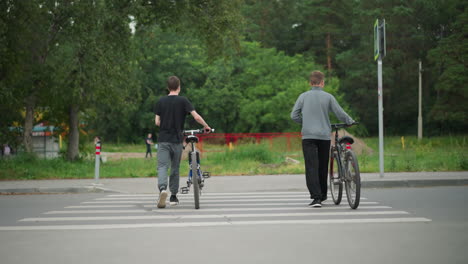 vista trasera de dos hermanos caminando uno al lado del otro con sus bicicletas a través de un cruce de peatones, el fondo presenta personas borrosas caminando, árboles verdes y una valla roja brillante