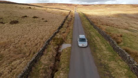 aerial moving drone shot of silver car driving down remote moorland road in english countryside on summer day