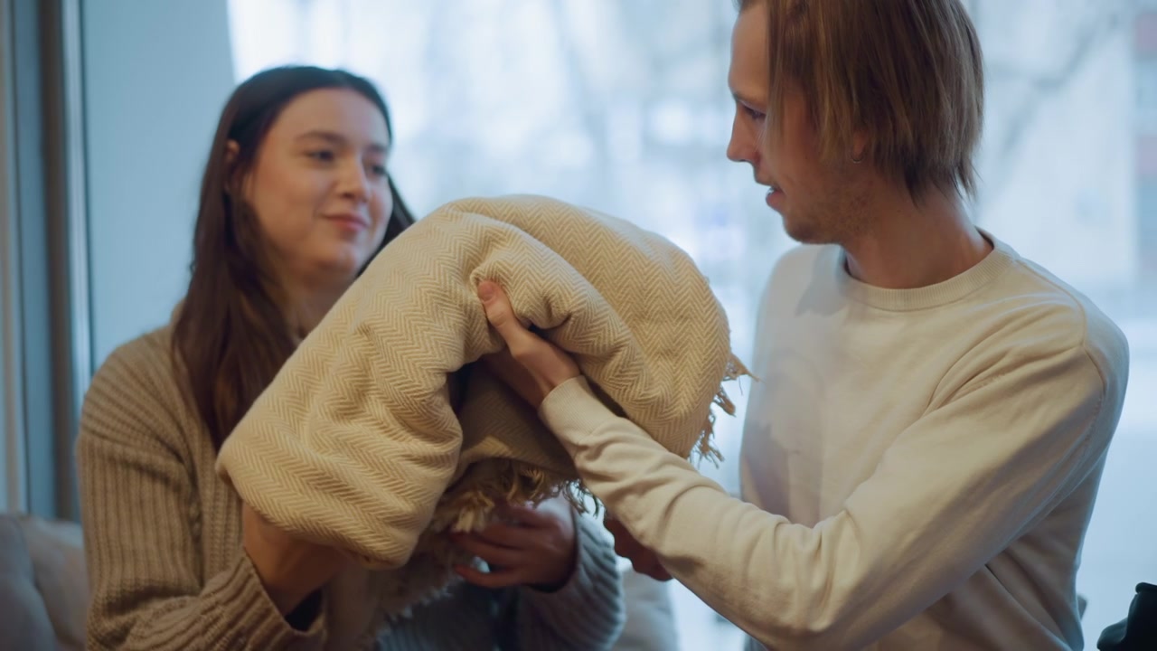 Close up of man and woman interacting with beige textured blanket indoors. Man hands over folded fabric to seated lady while they converse suggesting gift or exchange in soft lighting
