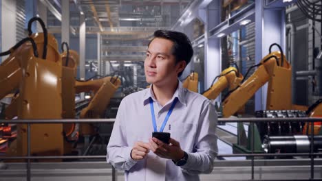 asian male professional worker standing with his smartphone with industrial robotic arms working, he observes by looking around before he come to concentrating with his smartphone