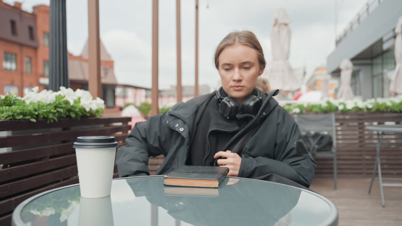 Ung, hvid kvinde ved cafébord udendørs; sidder med kaffekop og notesbøger, tager jakke på, justerer hovedtelefoner, lægger telefon ved siden af notesbog, betragter regnvåd skyline, stemningen er eftertænksom, scene urban