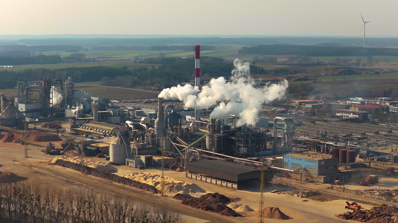 Panoramic aerial view of a large wood processing factory complex. Thick white smoke and steam rising from the chimneys, polluting the air