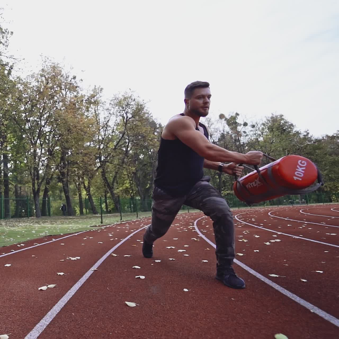 Strong athlete doing his workout on the stadium. Handsome muscular bodybuilder doing squats with weight outside. Slow motion.