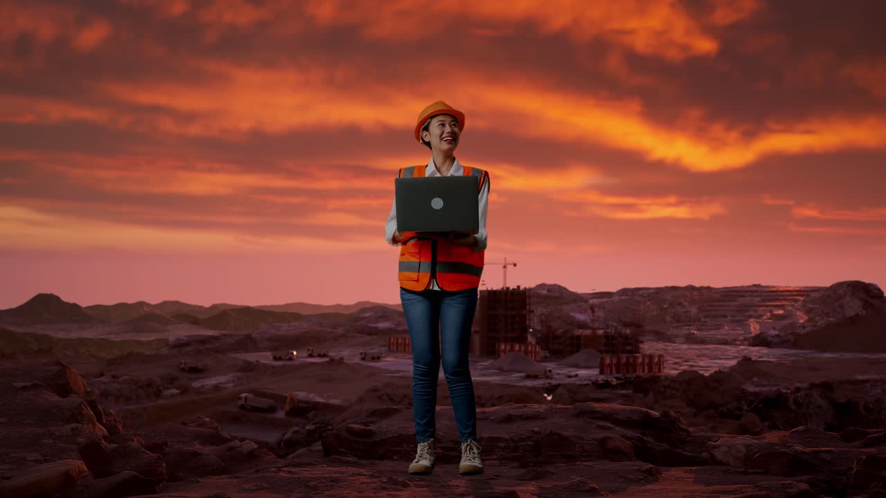 cuerpo lleno de ingeniera asiática con casco de seguridad trabajando en una computadora portátil y mirando a su alrededor mientras está de pie en el sitio de construcción, maquinaria de minería
