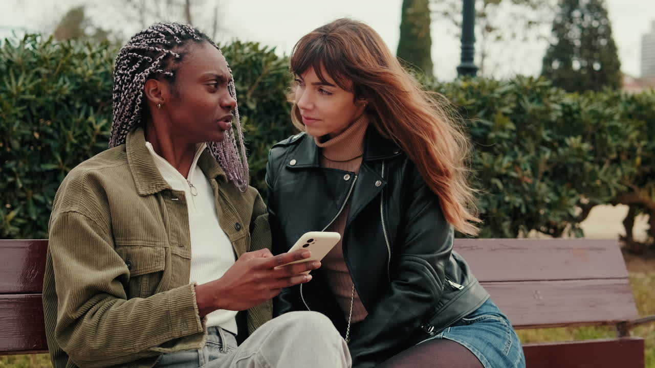 Two Young Women Talking and Looking at a Smartphone on a Park Bench