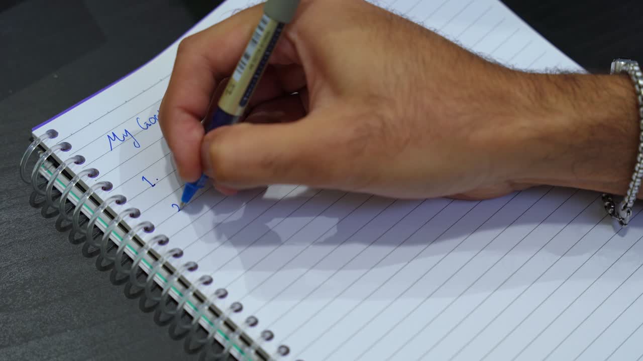 Close-up of hand writing “My Goals” in blue ink on spiral notebook. Ideal for motivation, productivity, planning, business strategy, or personal growth videos.