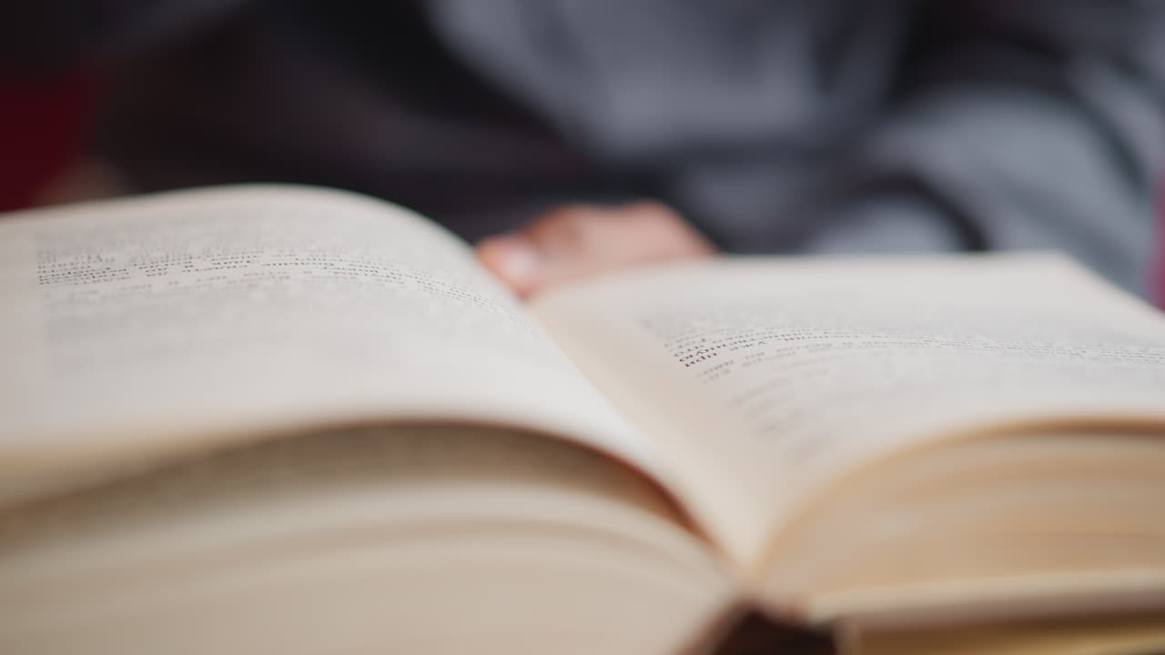 Close-up view of open book held by male turning page, blurred hand and shirt in background, sharp focus on printed text in dim indoor lighting, capturing moment of quiet reading or concentrated study