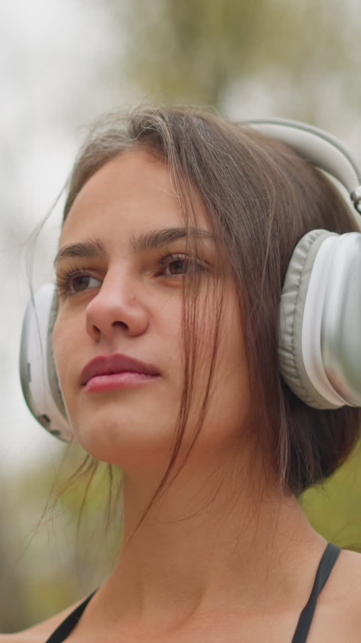 Close-up shot of beautiful young lady wearing headphones with focused expression while adjusting them on her head, she stands outdoors, blending into nature with a peaceful and concentrated demeanor