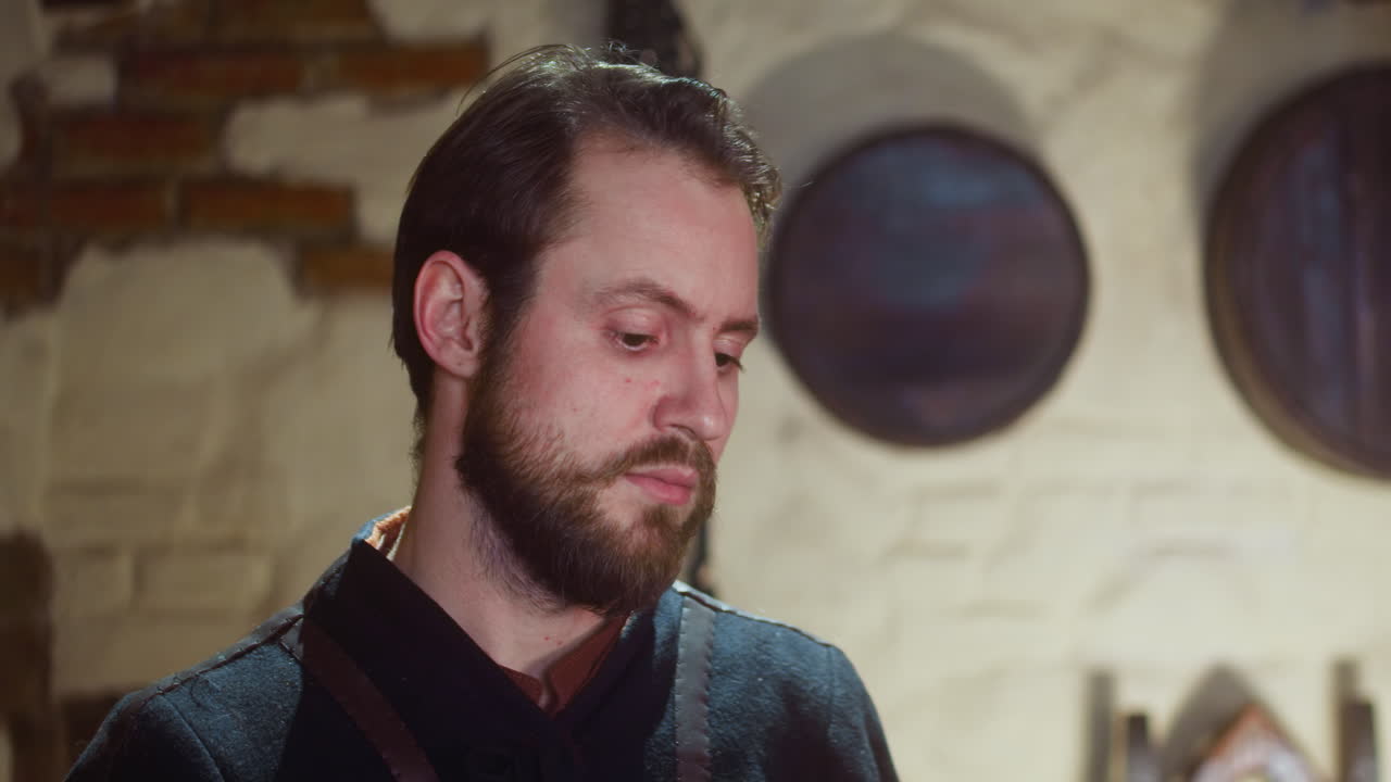 el hombre barbudo pasa la noche en una taberna medieval en primer plano. el huésped se inclina hacia adelante para tomar el artículo y camina en la antigua posada. fantasía oscura y participante del juego de rol