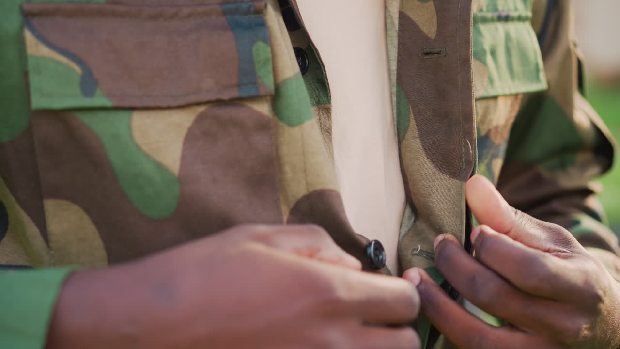 Black Soldier Buttoning Camo Jacket Outdoors, CloseUp Hands Fastening Buttons With Calm Determination And Soft Sunlight Tactile Ritual Conveys Resilience, Duty And Quiet Anticipation Of Return