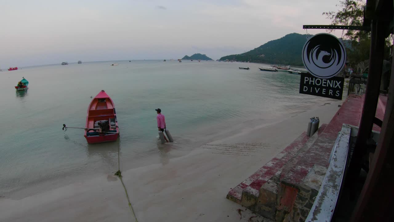 Scuba tanks are loaded onto a long narrow boat for the morning dives. Man carrying tanks from the beach in front of a dive shop. Morning light on a peaceful beach in Koh Tao, Thailand.