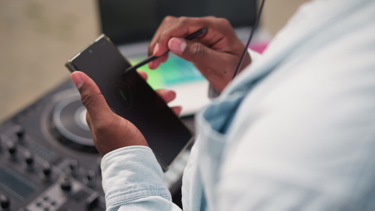 Sort DJ trykker på smartphone-pen nær mixer. Nærbillede af hænder, der justerer spilleliste, cue'er numre og forhåndsviser samples på touchskærmen. Ærme fra denimjakke. Koncentreret, urban studiestemning.