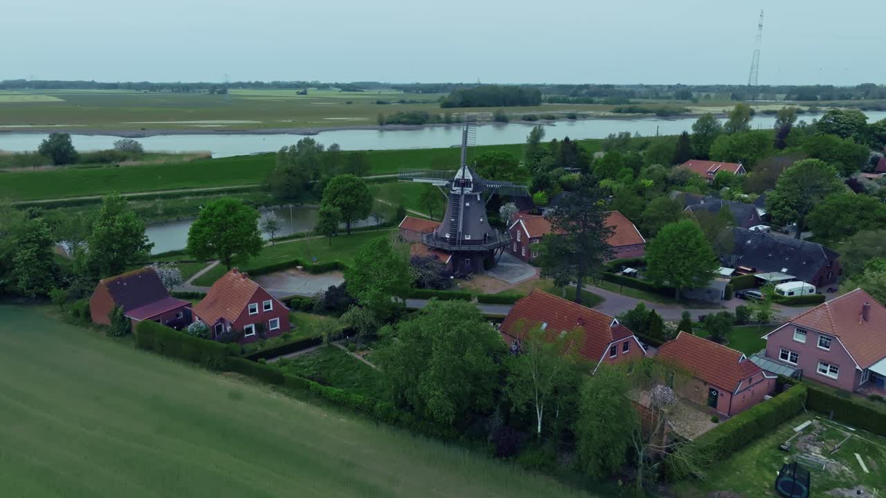 Drone slowly zooms in towards a windmill in a quiet green village. Lighting is natural with slightly cloudy sky.