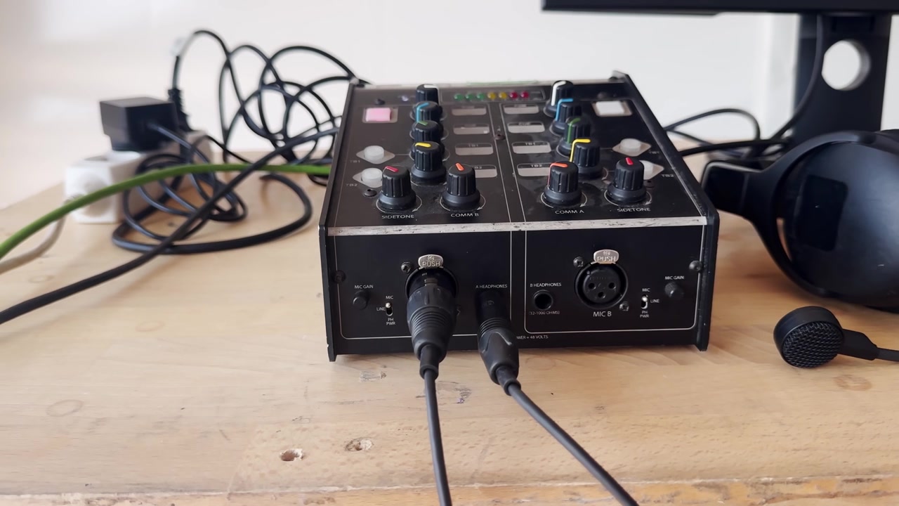 Unidad de intercomunicación de transmisión con cables conectados y micrófono de diadema sobre una mesa de madera, mostrando equipo de comunicación para producción en vivo y configuración de control de audio