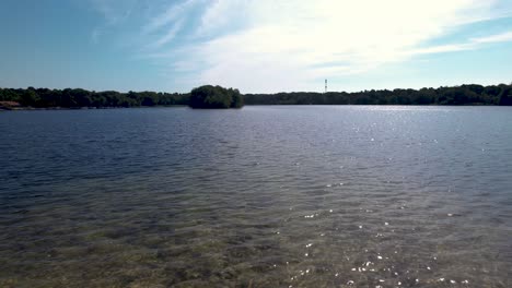 Constante-Movimiento-Aéreo-Hacia-Atrás-Desde-Arriba-Del-Agua-De-La-Laguna-Que-Revela-El-Camino-En-La-Península-En-El-Lago-De-Ijzeren-Man-Con-Juego-De-Sombras-Y-Follaje-Verde-A-Lo-Largo-Del-Camino-Y-La-Isla-En-El-Fondo