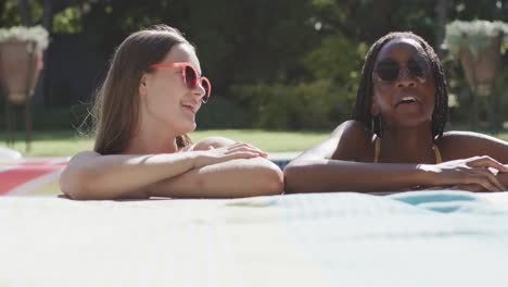 Two-happy-diverse-teenage-female-friends-in-sunglasses-talking-in-swimming-pool-in-slow-motion