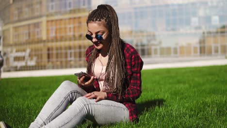 young woman with dreads relaxing in park sitting on the grass and listening to music. hipster girl in sunglasses moving her body