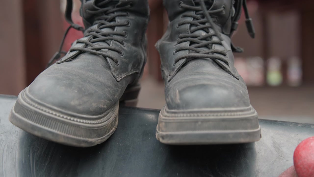 Closeup boots balancing on skateboard deck, worn leather laces and scuffed soles, red wheel peeking, urban park railing blurred, skater preparing for trick, tense preride mood with cinematic shallow