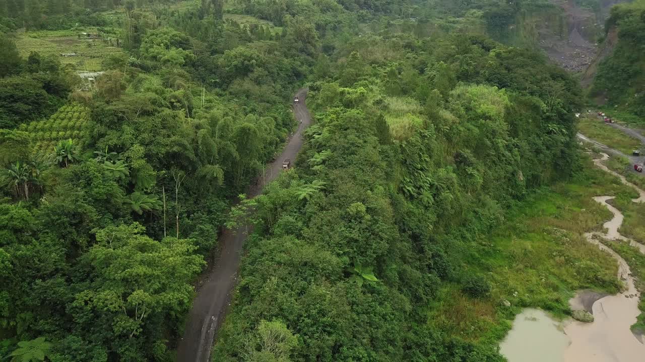 toma aérea de pájaros de camiones que circulan por caminos rurales entre bosques en la ladera del volcán merapi - transporte para mina de arena en indonesia - tiro ascendente