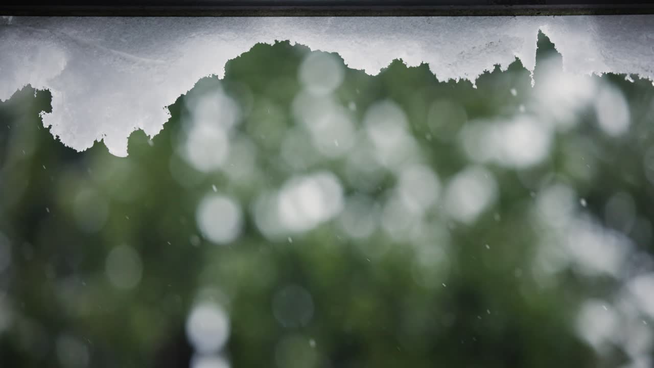 Snow hanging from a roof while it's snowing in the background with trees out of focus