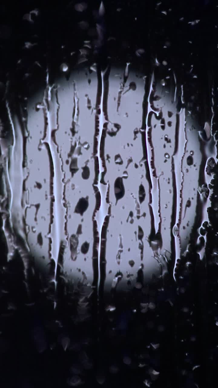 Circular Light ( Can Resemble The Moon ) behind window covered with sleet and raindrops streaming down the window at night. 9:16 Vertical