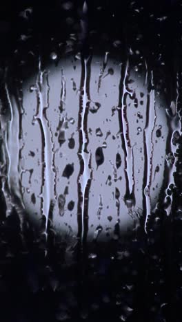 circular light ( can resemble the moon ) behind window covered with sleet and raindrops streaming down the window at night. 9:16 vertical