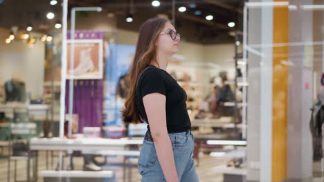 vista lateral de una mujer con camiseta negra y vaqueros caminando por delante de una elegante tienda minorista en un centro comercial, la fachada de vidrio refleja una iluminación brillante, con estantes llenos de accesorios de moda