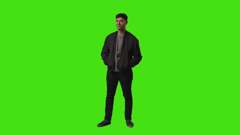 toma completa de un joven vestido casualmente sonriente de pie contra la pantalla verde poniendo las manos en los bolsillos