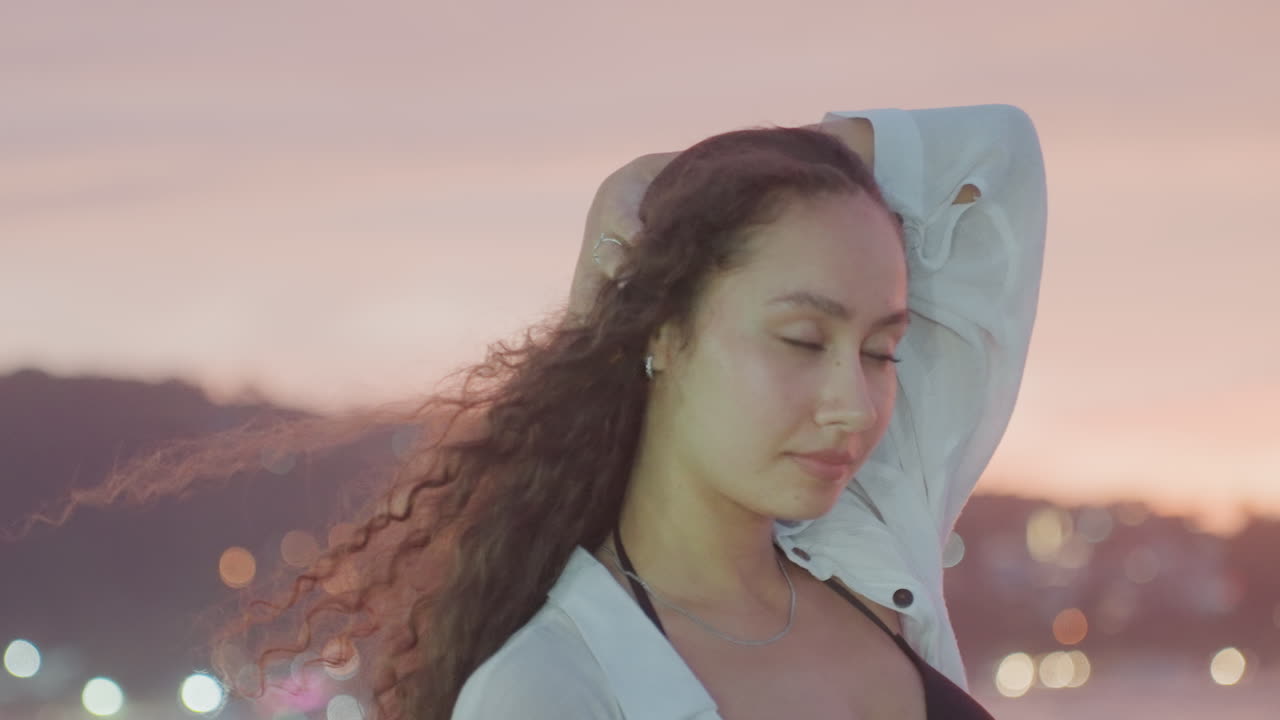 Woman gazes through hair by tranquil shoreline. Quiet dusk scene with city lights shimmering behind woman. Gentle evening setting showing woman with flowing hair by peaceful ocean under pastel sky
