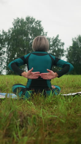 vista trasera de una mujer sentada en una alfombra de yoga en la postura de sukhasana practicando yoga con los ojos cerrados, abrazando la calma en un vasto campo de hierba bajo un cielo nublado, con árboles alineados en el fondo