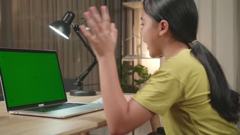 back view of asian kid girl sitting in a wheelchair while video call on laptop computer with mock up green screen at home