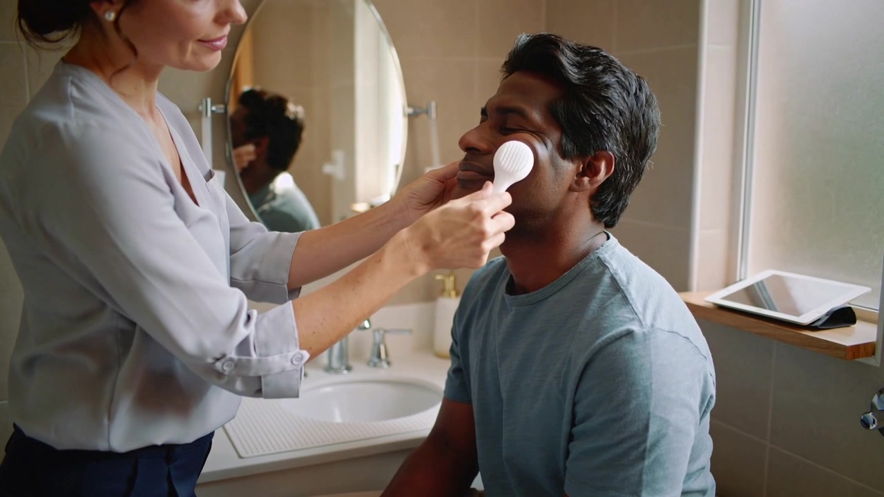 Bringing facial brush, woman gently cleansing seated man's cheek for care in bathroom, tablet near