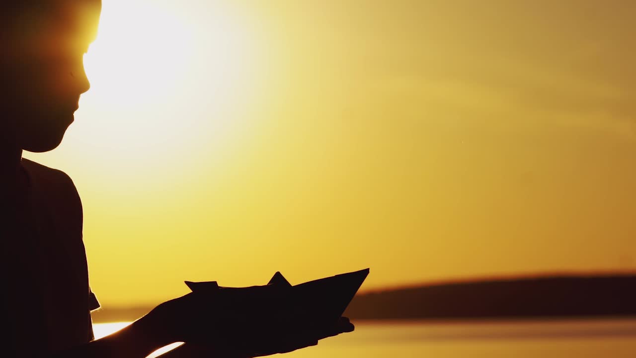 boy is playing with origami of handmade in the form of a boat on the background of sunset by the river in the summer