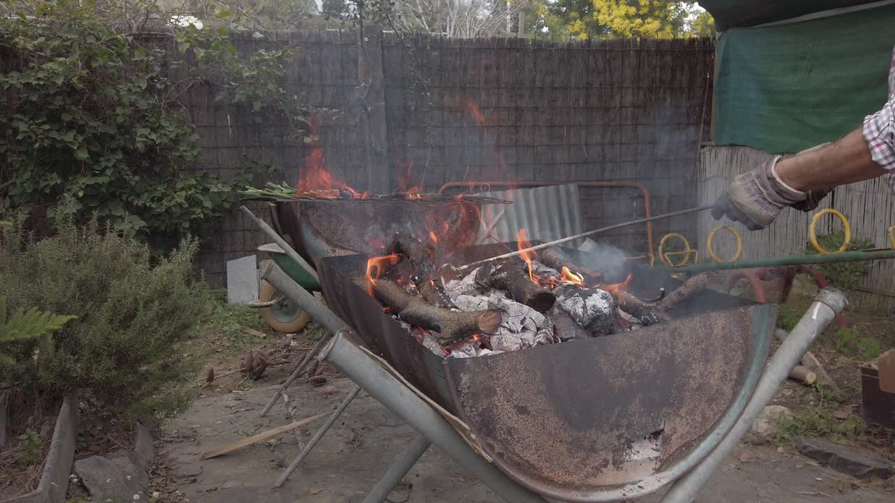 Professional chef stoking wood fire in metal barbecue grill, creating flames and heat while preparing outdoor meal in lush garden setting