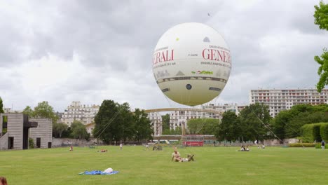 a large generali tethered balloon floating above parc andré-citroën in paris, with people relaxing on the grass and city buildings in the background.