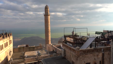 as the camera pans to the right, we see the roof cafes of mardin and the minaret of the great mosque behind them, and the magnificent view of mesopotamia at the back