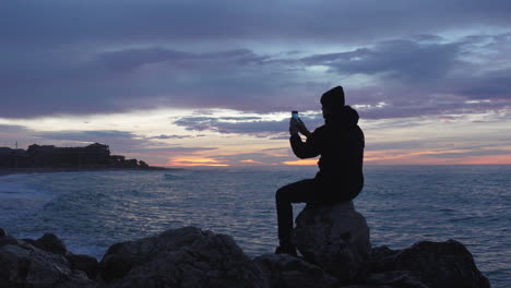 blue-hour-at-sea,-man-sits-on-a-rock-enjoys-his-time-looking-at-sunset-beach,-takes-photo-and-leaves