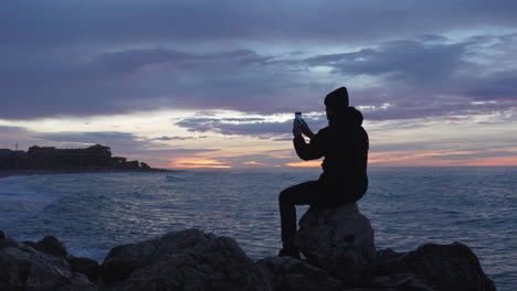 Blaue-Stunde-Auf-See,-Mann-Sitzt-Auf-Einem-Felsen,-Genießt-Seine-Zeit-Mit-Blick-Auf-Den-Sonnenuntergangsstrand,-Macht-Ein-Foto-Und-Geht