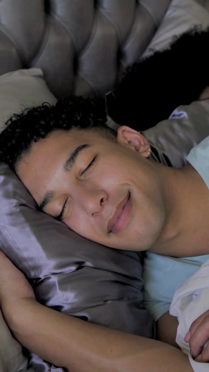 Vertikal video: Kameraet trækker sig tilbage, mand i blå top sover og smiler på en satinpude, med plads til tekst.