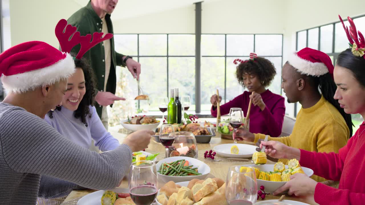 Diverse friends passing vegetables and rolls at table prompted by host carving chicken for holiday