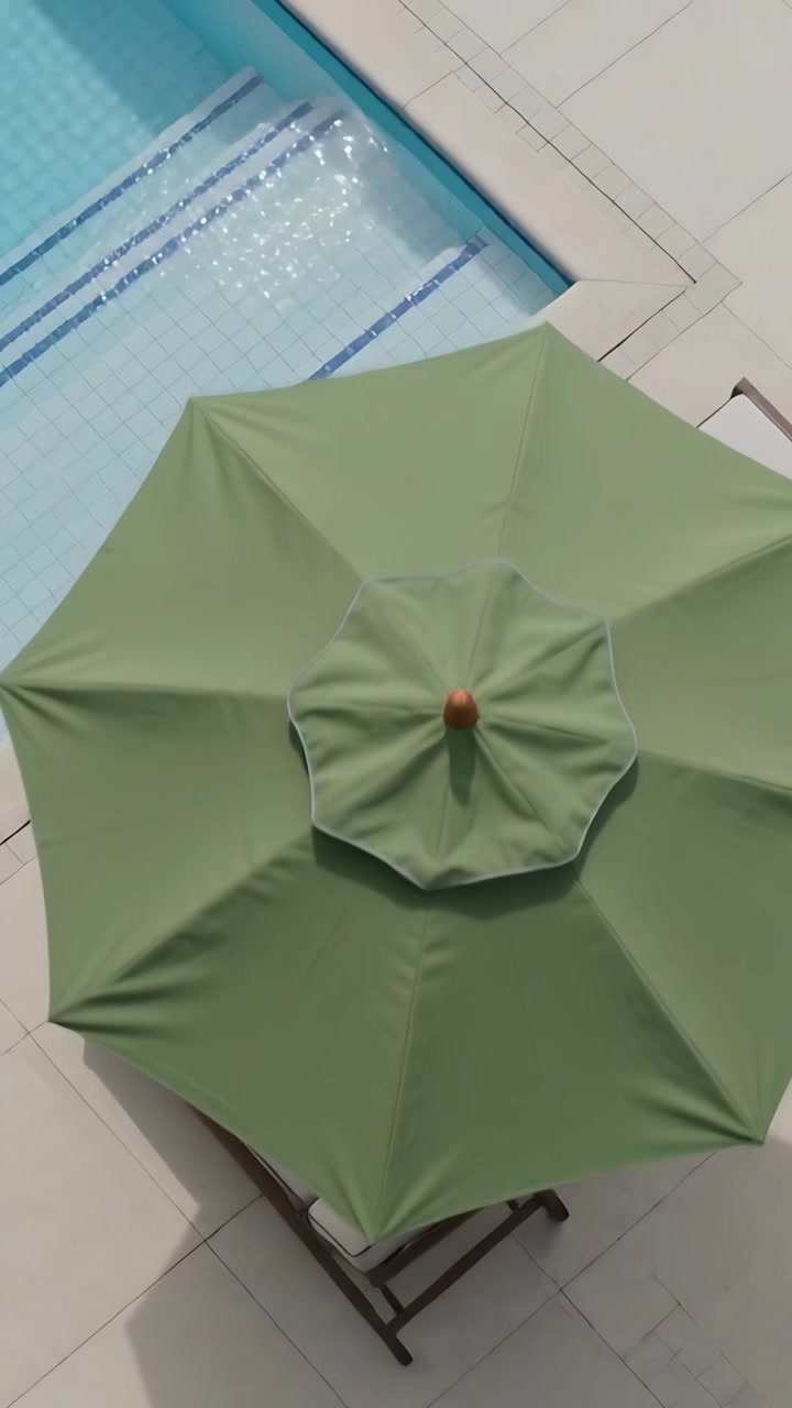 Video verticale: Brezza che muove le fronde delle palme, la telecamera si sposta per rivelare un ombrellone verde sopra una piscina.