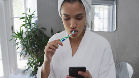 mujer de raza mixta cepillándose los dientes y usando su teléfono inteligente en el baño
