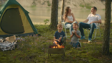A-family-in-nature-parents-watch-as-two-boys-at-the-fire-roast-marshmallows-on-sticks-in-the-background-of-the-tent.-Tent-camp-as-a-family