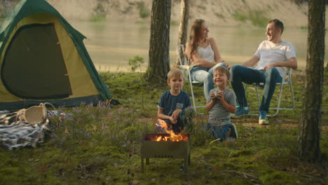 a family in nature parents watch as two boys at the fire roast marshmallows on sticks in the background of the tent. tent camp as a family