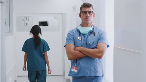 Portrait-Of-Mature-Male-Doctor-Wearing-Scrubs-Standing-In-Busy-Hospital-Corridor